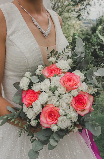 A bride wearing a sleeveless, ivory wedding dress with subtle sparkle details and a diamond necklace, holding a large floral bouquet outdoors with lush green trees in the background. The bouquet featu