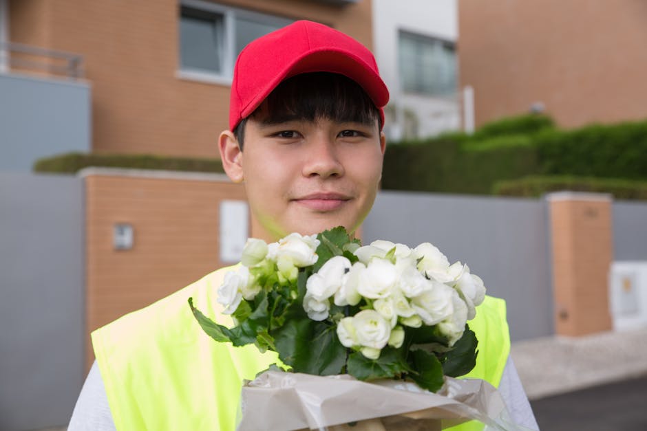 A young delivery person wearing a bright yellow safety vest and a red cap stands outside a modern residential building, holding a bouquet of fresh white flowers wrapped in white paper. The flowers app