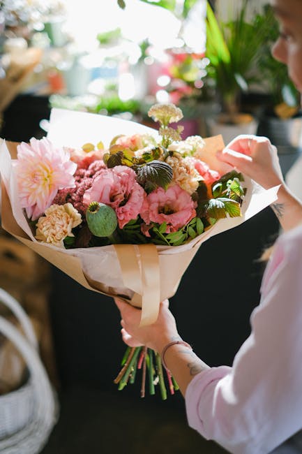 A person holding a large, vibrant floral bouquet featuring pink peonies, cream roses, and white carnations, accented with green foliage and wrapped in beige paper with a satin ribbon. The bouquet is p