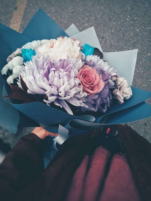 A hand holding a bouquet of fresh flowers arranged with a large, soft pink rose, white chrysanthemums, purple statice, and small blue accent flowers, wrapped in elegant blue and white paper with a glo
