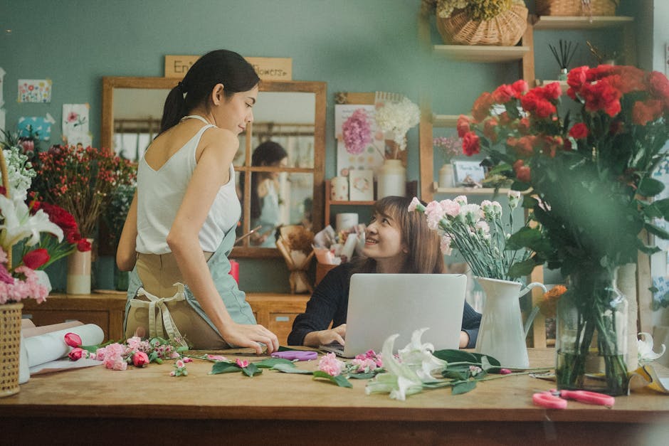 A young woman in a floral shop, smiling and engaging with a customer at the counter. She is wearing a light-colored sleeveless top and a beige apron tied at the waist. The customer, a woman with long